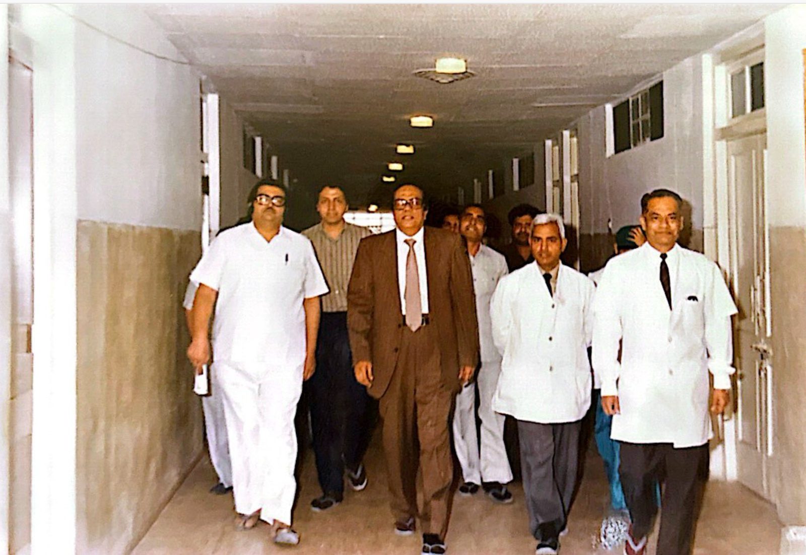 Dr. J.P. Sethi in a brown suit walking a hospital corridor with colleagues in white coats